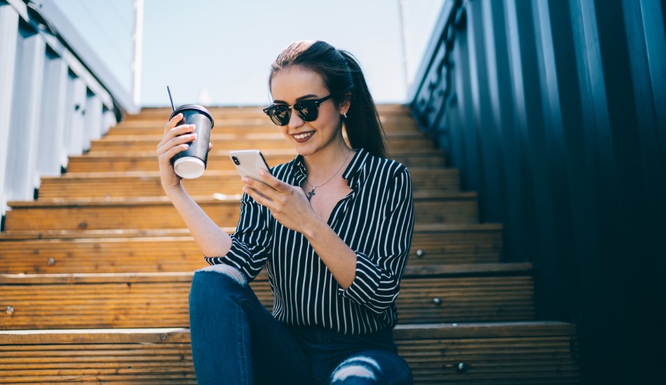 Woman sitting on stairs drinking coffee while smiling at her phone